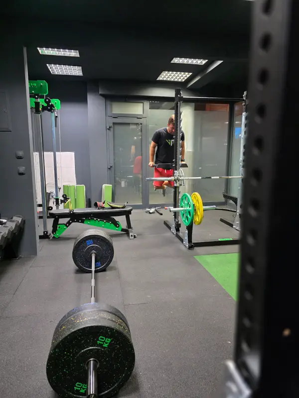 Pogled na dvoranu — trener u V-Lift majici trenira kraj stalka i zelene klupe
